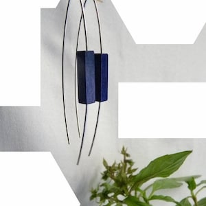 Op de afbeelding: Een paar moderne oorbellen met lange, zilverkleurige draden met een gebogen ontwerp. Elke oorbel heeft een donkerblauwe, rechthoekige houten accent. De oorbellen worden gepresenteerd tegen een witte achtergrond, met een takje groene bladeren.