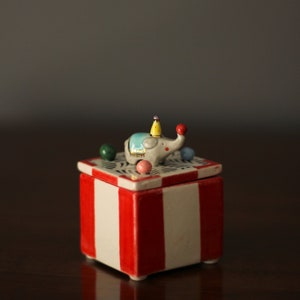 Könnte beinhalten: Keramik-Schmuckkästchen mit rot-weiß gestreiftem Sockel. Der Deckel zeigt einen grauen Elefanten mit blauer Decke, gelbem Hut und rotem Ball auf seinem Rüssel. Bunte Kugeln umgeben den Elefanten.