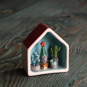 May include: A ceramic house-shaped planter with a red roof and a light blue interior. It contains miniature cacti in shades of green and blue, each in a small pot. The planter is set on a teal wooden surface.