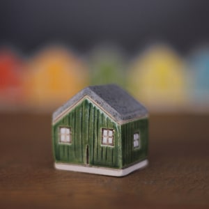 May include: A small green ceramic house with white windows and a gray roof. The house is sitting on a wooden surface.