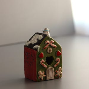 以下が含まれることがあります： セラミック製のクリスマスハウスの置物。家は、赤いテクスチャ加工された側面と、キャンディケイン、ジンジャーブレッドマン、ハートで飾られた緑色の前面を備えています。白い雪で覆われた屋根には雪だるまが座っています。中央にはハート型の開口部があります。