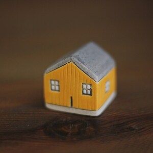 May include: A small, yellow ceramic house with a grey roof and two windows. The house is sitting on a wooden surface.