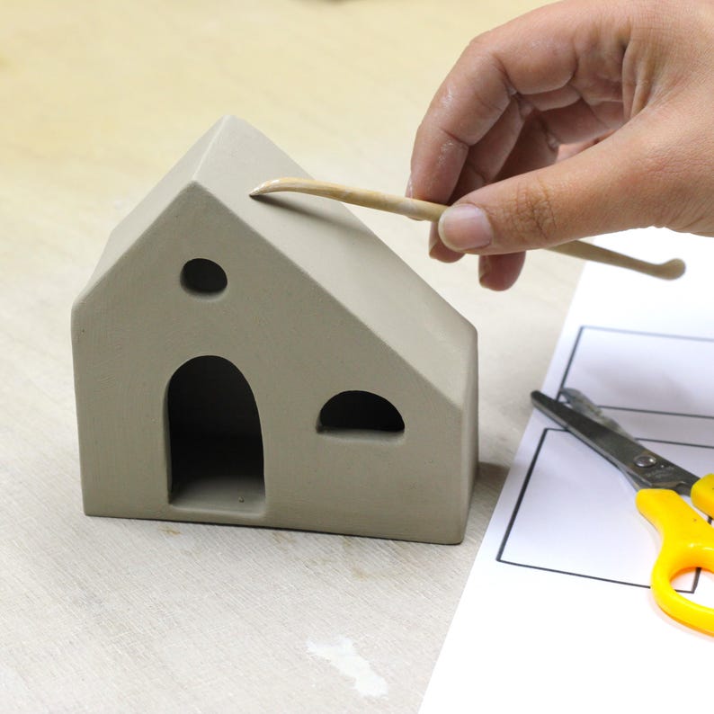 May include: A person uses a small wooden tool to smooth the top of a gray clay house. The house has three openings, one large and two small. A pair of yellow-handled scissors is on a white sheet of paper with black lines.