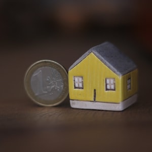 May include: A small yellow house figurine with a gray roof and white trim sits next to a one euro coin.