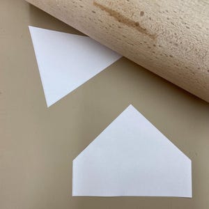 May include: Two white paper shapes, one triangle and one home plate shape, on a light brown surface. A wooden rolling pin is partially visible in the top left corner of the image.