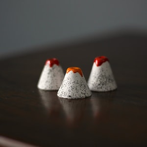 May include: Three small ceramic volcano-shaped figurines with white glaze and black speckles. The tops of the volcanoes are painted in red, orange, and yellow.