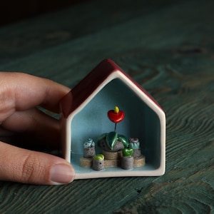 May include: A miniature ceramic house with a red roof and light blue interior. Inside, a small plant arrangement features a red flower and several potted plants. The piece is held by a hand, showcasing the intricate details of the handcrafted item.