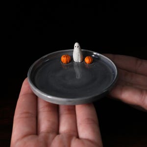 Peut inclure: Un petit plat en céramique gris avec un fantôme blanc et deux citrouilles orange sur le dessus. Le plat est dans la paume d'une main.