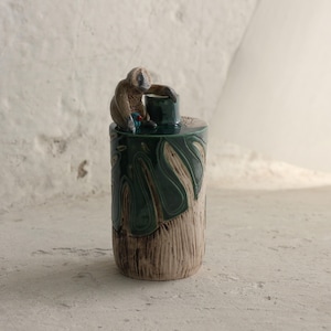 Puede incluir: Un jarrón decorativo de cerámica con una figura de mono. El jarrón tiene forma cilíndrica con un diseño texturizado verde y marrón. El mono marrón está sentado encima del jarrón. El diseño sugiere un artículo hecho a mano, con una estética artística y única.