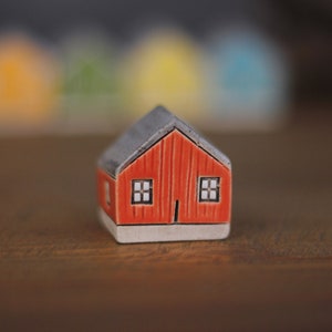 May include: A small red ceramic house with a grey roof and two white windows. The house is sitting on a wooden surface.
