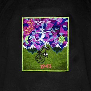 May include: Black t-shirt featuring a colorful graphic with the text "Bicycle Day" and the year "1943." The graphic depicts a person riding a bicycle with a surreal background of mountains, flowers, and a sun.