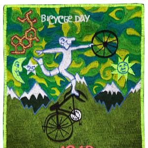 Puede incluir: Un parche bordado verde con una figura blanca montando una bicicleta con una gran rueda delantera. El parche tiene un fondo verde, amarillo y azul con un sol, una luna y estrellas. El texto "BICYCLE DAY 1943" está bordado en el parche.
