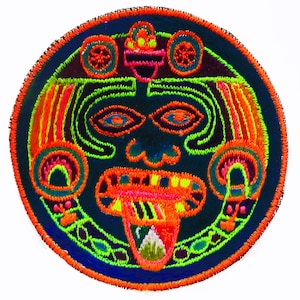 Peut inclure: Écusson brodé circulaire représentant un visage stylisé. Le design utilise des fils orange fluo, verts et bleus sur un fond bleu foncé. Le motif présente une bordure détaillée et rappelle l'art ancien.