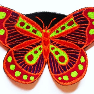 Pode incluir: Patch de borboleta bordada com base preta. A borboleta apresenta cores vibrantes: asas vermelhas com detalhes verdes e roxos e contorno laranja. O design é detalhado com padrões costurados, criando uma aparência texturizada.