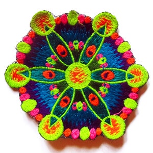Könnte beinhalten: Gestickter Aufnäher mit einem symmetrischen Design. Der Aufnäher zeigt eine zentrale blütenartige Form mit strahlenförmigen Blütenblättern in Neongrün, Orange und Pink, vor einem Hintergrund aus blauen und violetten Fäden. Der Aufnäher befindet sich auf weißem Hintergrund.