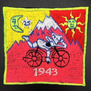 May include: A red, yellow, and white embroidered patch with a cartoon figure riding a bicycle up a mountain. The patch has a crescent moon, a star, and a sun with a face. The text "1943" is at the bottom of the patch.