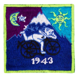 Peut inclure: Un patch brodé bleu, vert et violet avec une scène de montagne blanche. Une personne fait du vélo en montant la montagne. Le soleil et la lune sont dans le ciel. L'année 1943 est brodée en blanc sur le patch.