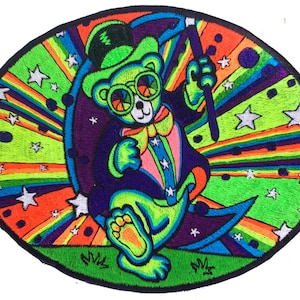 Peut inclure: Écusson brodé ovale représentant un ours coloré portant un chapeau haut de forme, des lunettes et un smoking. L'ours tient une baguette magique sur un fond d'étoiles et de rayures arc-en-ciel. Bordure noire.