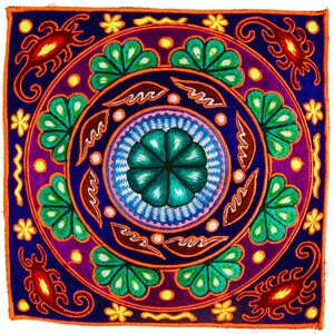 Pode incluir: Um tecido quadrado bordado com cores vibrantes com um desenho circular central que apresenta uma flor verde, azul e rosa. A borda é decorada com escorpiões estilizados e motivos florais.