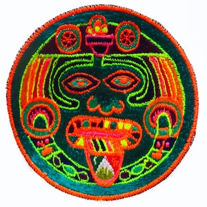 Pode incluir: Um patch redondo bordado com um rosto estilizado em cores vibrantes. O design inclui um fundo turquesa, detalhes em laranja e verde néon e uma borda vermelha. O rosto tem olhos grandes, uma boca larga e um cocar detalhado.