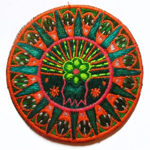 Peut inclure: Un patch rond brodé avec un motif floral vert, rose et orange. Le motif est entouré d'une bordure verte et orange.