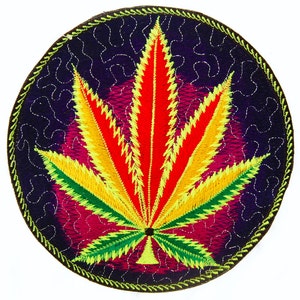 May include: Circular embroidered patch with a cannabis leaf design. The leaf is depicted in red, yellow, and green, set against a dark purple background with a decorative stitch pattern. The patch has a green border, suitable for clothing or accessories.