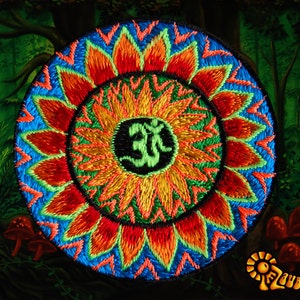 May include: A vibrant, embroidered mandala with a central Om symbol. The design features a circular pattern with red, orange, yellow, and blue threads. The background depicts a forest scene with mushrooms and the text "Zauberwald".