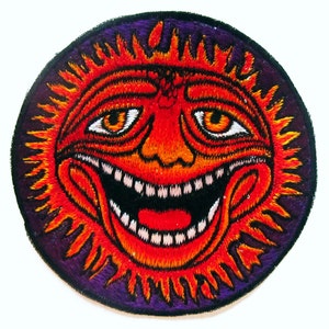 Puede incluir: Parche bordado circular que representa un sol sonriente con rayos naranjas y una cara roja. El sol tiene una amplia sonrisa, dientes blancos y ojos naranjas, sobre un borde morado.