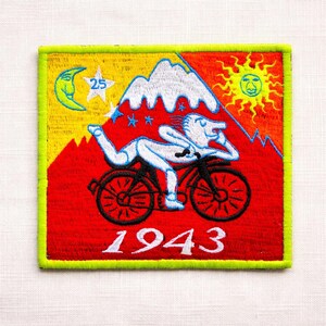 May include: Embroidered patch with a red background, featuring a white figure on a bicycle, mountains, a sun, moon, and stars. The year "1943" is displayed in white. The patch has a yellow border with a green outline.