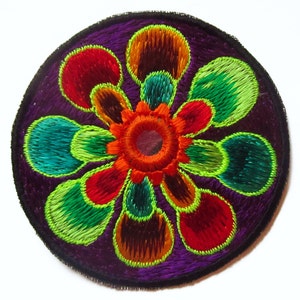 May include: A round, embroidered patch with a floral design. The patch features a deep purple background with a black border. The flower design includes red, orange, green, and teal petals, with a central orange circle.