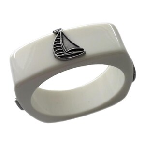 May include: A square-shaped white bangle bracelet featuring a small, metallic silver-toned sailboat charm attached to the top surface. The bracelet has a smooth, glossy finish and a rounded interior opening, showcasing a clean and simple design.