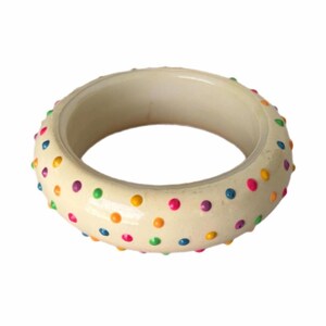May include: Ivory-colored bangle bracelet with a glossy finish. The bracelet is adorned with evenly spaced, raised, colorful polka dots in shades of pink, purple, blue, green, and yellow. The bracelet is a circular shape.