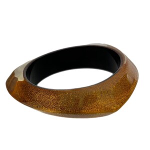 May include: A brown and gold bangle bracelet with a black interior. The bracelet is made of a shiny, textured material.