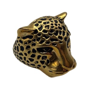May include: A gold-toned ring shaped like a leopard head, featuring a metallic finish with recessed black spots across the surface. The design captures the feline facial features, including pointed ears, defined eyes, and a snout, in a solid metal form.