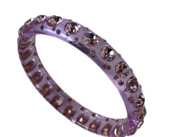 May include: A transparent lavender bracelet adorned with numerous pink gemstones. The bracelet has a rounded shape and is set against a white background. The gemstones are evenly spaced around the band, adding sparkle and detail.
