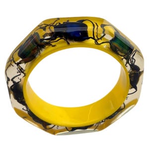 May include: A unique, octagonal bangle bracelet. The clear resin is embedded with several beetles, showcasing iridescent green and blue hues. The interior of the bracelet is a bright yellow, creating a striking contrast. A distinctive piece of jewelry.