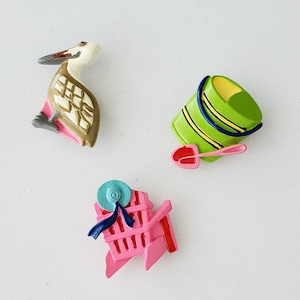 May include: A set of three colorful plastic figurines, including a pelican, a beach chair, and a bucket and shovel. The pelican is brown and white with a pink beak. The beach chair is pink with a blue and white striped canopy. The bucket and shovel are green with yellow stripes.