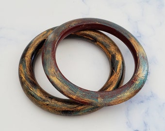 May include: Two wooden bangles with a layered, abstract design in shades of brown, gold, green, and red.
