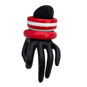 May include: A black hand-shaped sculpture supports red and white striped and solid red bangles, with a black oval shape on top. The sculpture is set against a white background.