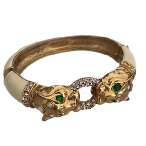 May include: Gold-tone hinged bracelet with cream enamel and two panther heads. The panther heads have green eyes and are embellished with clear crystals. A ring of crystals connects the two panther heads.