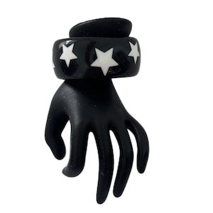 May include: A wide, glossy black bangle bracelet featuring a repeating pattern of solid white five-pointed stars encircling the exterior. The bracelet has a smooth, polished finish and a thick, rounded band construction.
