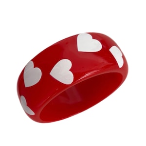 May include: A red bangle bracelet with white heart-shaped designs. The wide, glossy bracelet is a bold accessory, perfect for adding a pop of color and a touch of whimsy to any outfit. The bracelet's width is approximately 1 inch.
