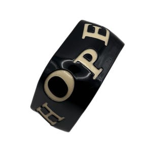 May include: A wide, glossy black ring featuring the word HOPE in raised, cream-colored capital letters. The band has a smooth, polished finish with the letters evenly spaced around the circumference of the ring, creating a bold, high-contrast look.