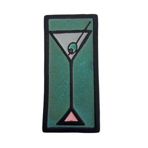 May include: Rectangular ceramic tile featuring a stylized martini glass with an olive. The glass is white with a black outline, set against a speckled teal background. A black border frames the design, with a pink triangle at the base.