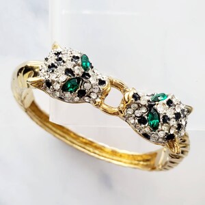 May include: A gold bangle bracelet with two panther heads on either side. The panther heads are adorned with black enamel, clear and green rhinestones.