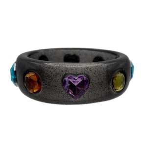 May include: A dark gray band ring featuring a textured finish and embedded faceted gemstones. The stones include a central purple heart-shaped gem flanked by a round orange gem and a round yellow-green gem, with blue gems visible on the sides.