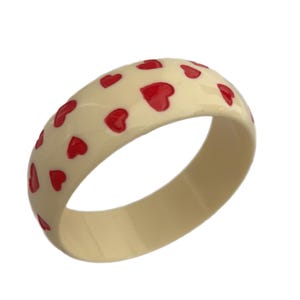 May include: Cream-colored bangle bracelet adorned with red heart-shaped designs. The bracelet has a smooth, glossy finish and a rounded shape. A charming accessory for everyday wear.