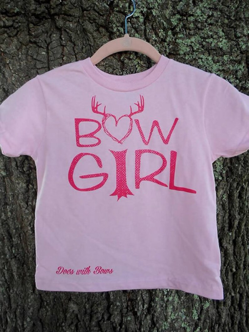 May include: Pink t-shirt with a red graphic of a heart with antlers and the text "BOW GIRL" and the text "Does with Bows" at the bottom.