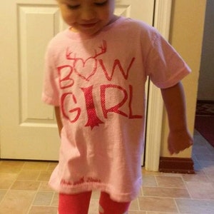 May include: A young girl wearing a pink shirt with the words "Bow Girl" in pink lettering and a pink heart. She is also wearing pink and white polka dot tights and pink shoes.