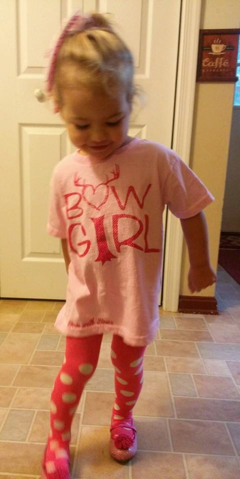 May include: A young girl wearing a pink shirt with the words "Bow Girl" in pink lettering and a pink heart. She is also wearing pink and white polka dot tights and pink shoes.